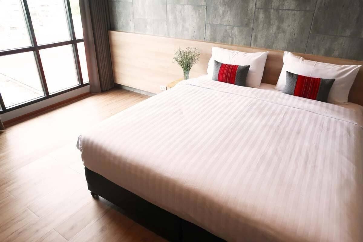 Una cama grande con una colcha de color claro en una habitación con pared de concreto y ventana.