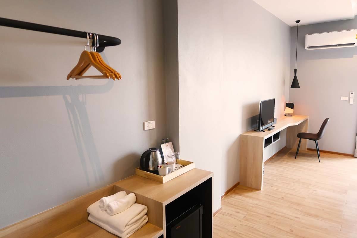 Une chambre moderne avec un bureau en bois, une télévision et une chaise. On y trouve une étagère avec des serviettes, ainsi qu'un plateau avec une bouilloire et des tasses.