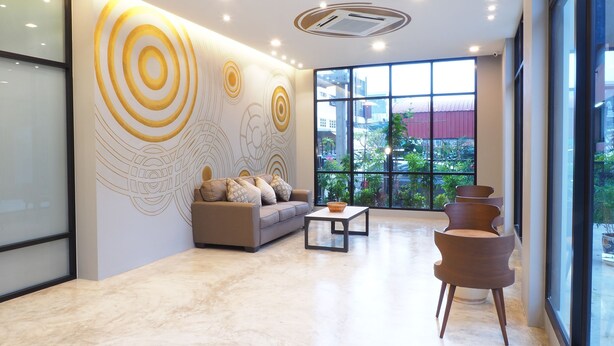 A modern lobby with a sofa, coffee table, and chairs, large windows with a view, and decorative wall art. - Sans Hotel (Ratchaburi)
