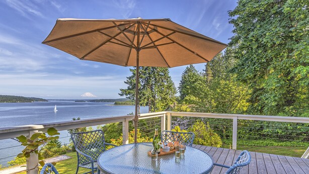 Une terrasse dotée d’une table et de chaises en verre, d’un parasol et offrant une vue magnifique sur un lac et des montagnes.
