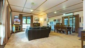 A spacious living room with a large black leather sofa, a dining area with wooden chairs, and a kitchen visible through an open doorway. - A lovely rustic cottage with all the modern conveniences (Gawler South)