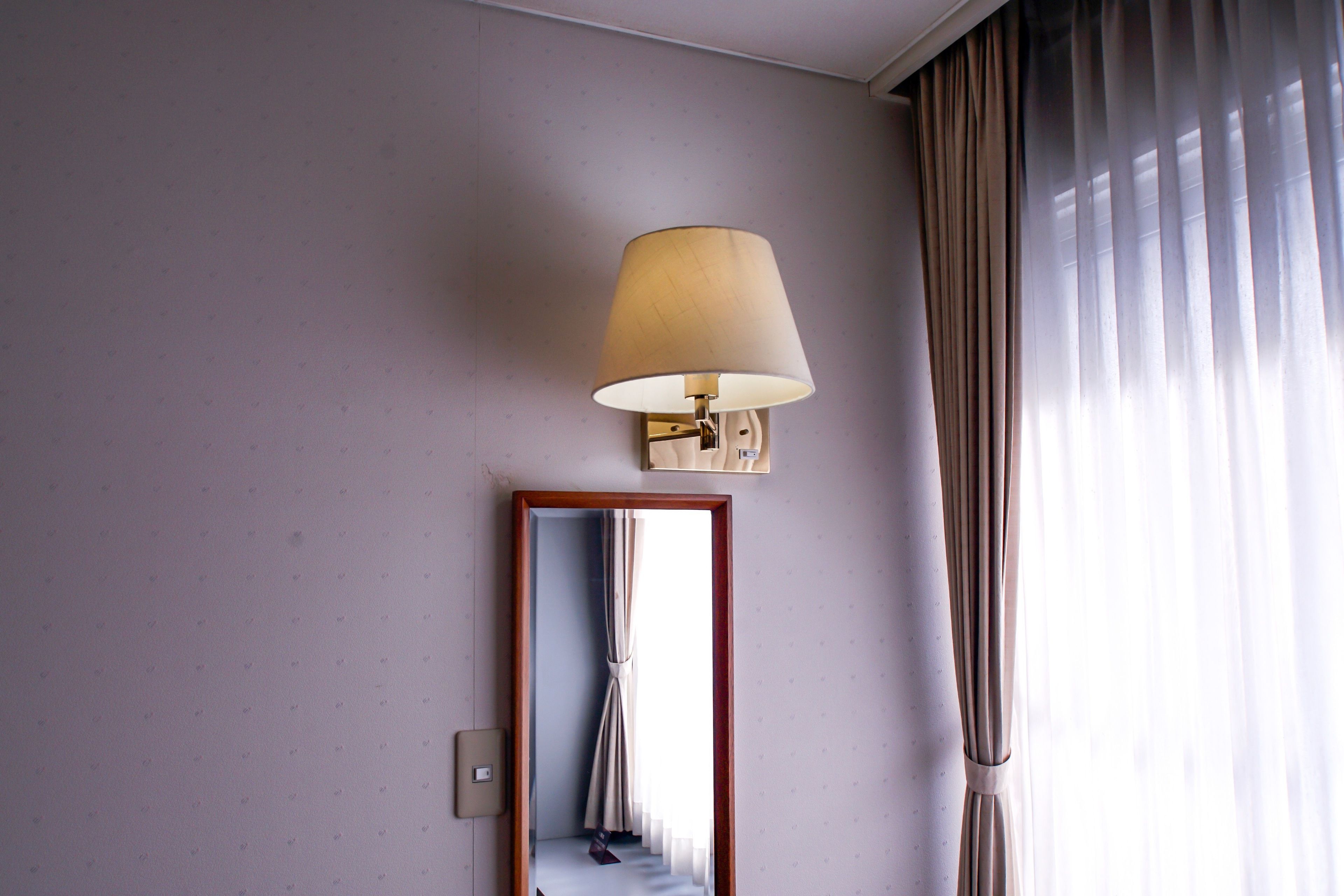 A hotel room with a wall-mounted lamp, a mirror, and a window with curtains.