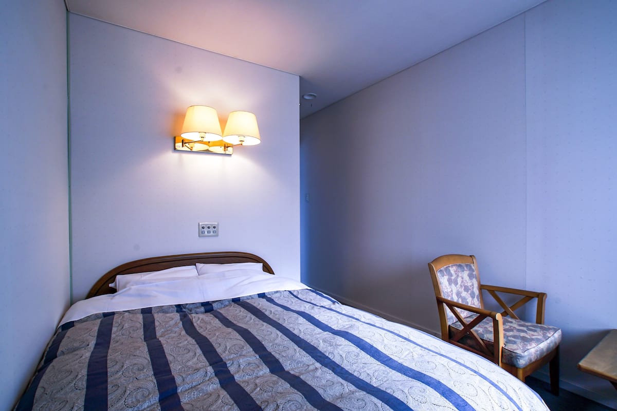 A neatly made bed with a patterned bedspread, a wall-mounted lamp, and a wooden chair.