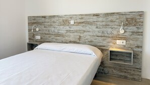 Un lit avec un matelas et des oreillers blancs, une tête de lit en bois dotée d’un éclairage intégré et une petite table de chevet.