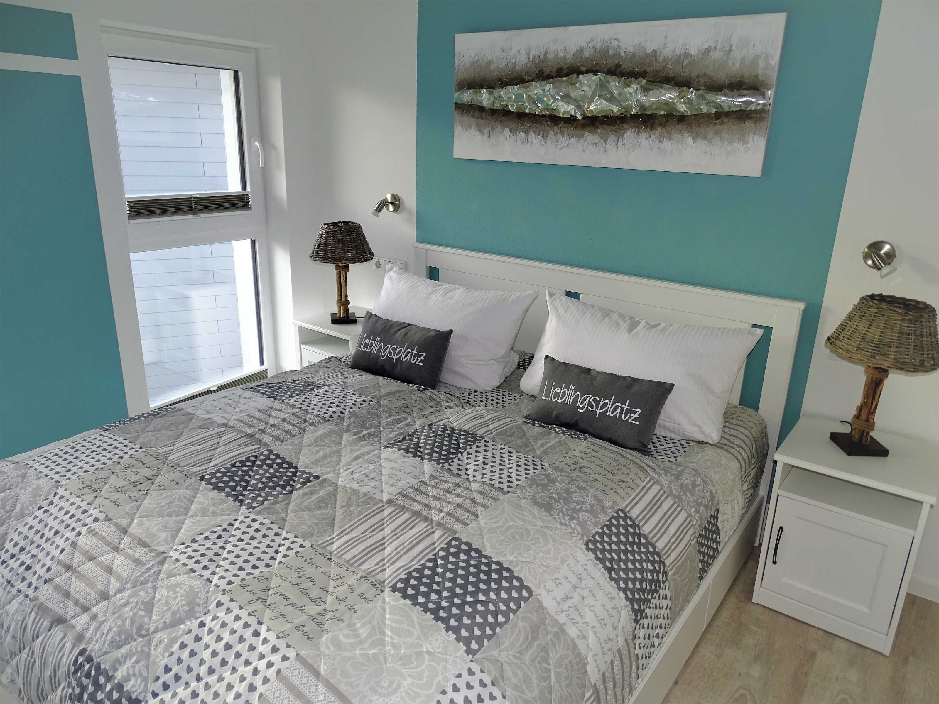 A neatly made bed with patterned bedding and pillows, a window with blinds, a wall-mounted lamp, and a painting above the bed.