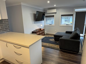 A modern kitchen with a countertop and cabinets, a flat-screen TV mounted on the wall, a sofa, and a window with a view of the outside. - Ulladulla Motel (Ulladulla)