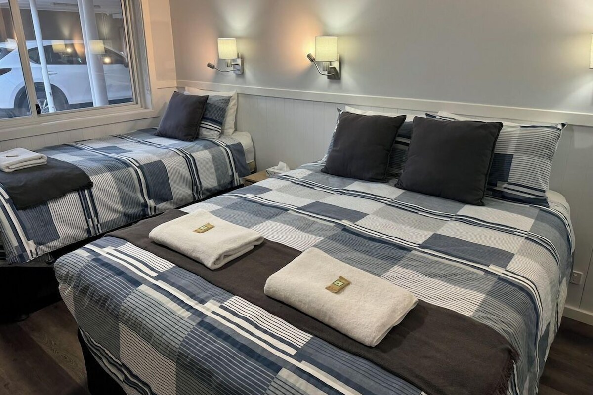 A hotel room with two beds, each with striped bedding and pillows, a window with a view of a white car outside, and wall-mounted lamps.