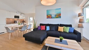 Un espai de vida modern amb un sofà seccional negre, una taula de dinar amb caires, una zona de cuina i un finestral gran que ofereix llum natural.