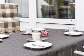 A dining table set with plates, cups, and a bowl, with a window and a plaid cushion in the background.