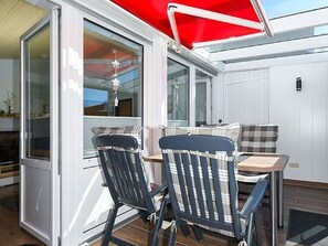Een overdekt terras met een tafel en stoelen, een glazen deur naar binnen en een rood zonnescherm.