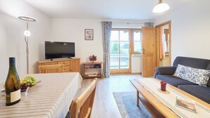 Een moderne woonkamer met een flatscreen televisie, houten meubels, een bank met opvallende kussens, een eettafel en een groot raam met gordijnen.