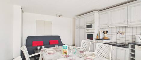 Un comedor con una mesa dispuesta para cuatro personas, sillas blancas, un sofá con cojines rojos, y una cocina con armarios y electrodomésticos blancos.