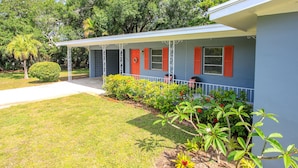 A single-story building with a covered porch, a small garden with various plants, and a clear sky.