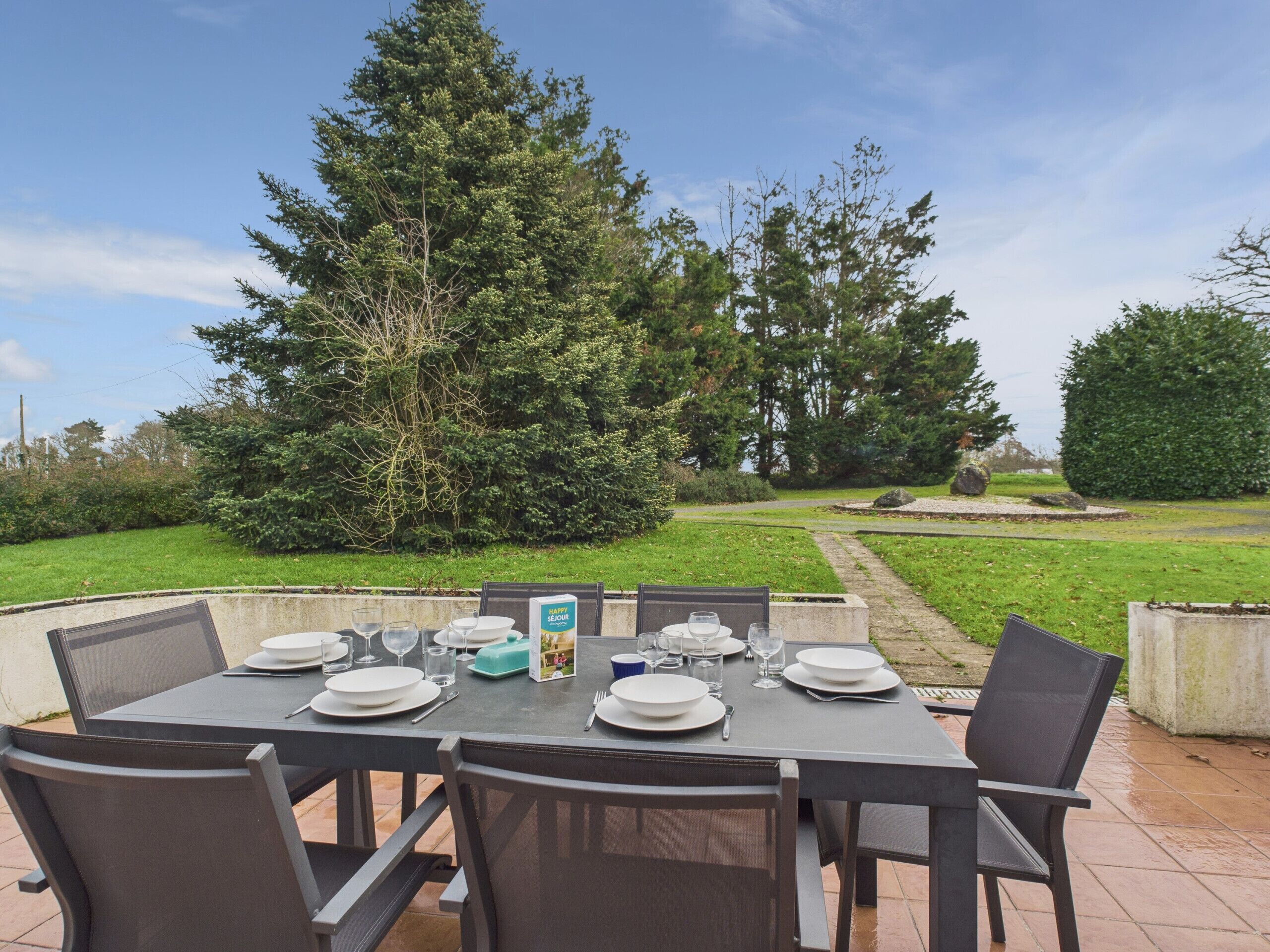 Un espace extérieur aménagé pour les repas, avec une table dressée pour six personnes, entourée de chaises, et un jardin et des arbres en arrière-plan.