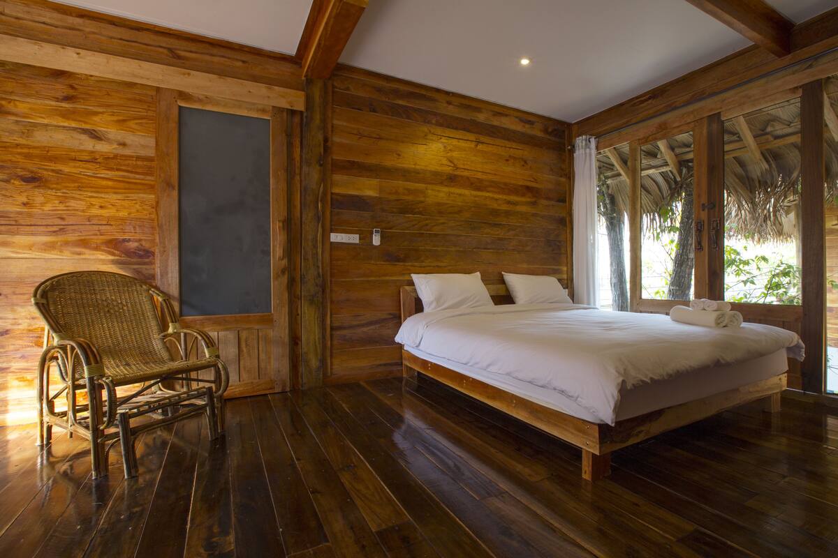 A wooden room with a bed, a wicker chair, and a large window.