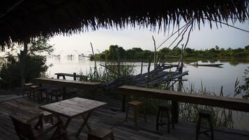 A wooden deck with tables and chairs overlooking a calm body of water, a dock extending into the water, and dense greenery in the background.