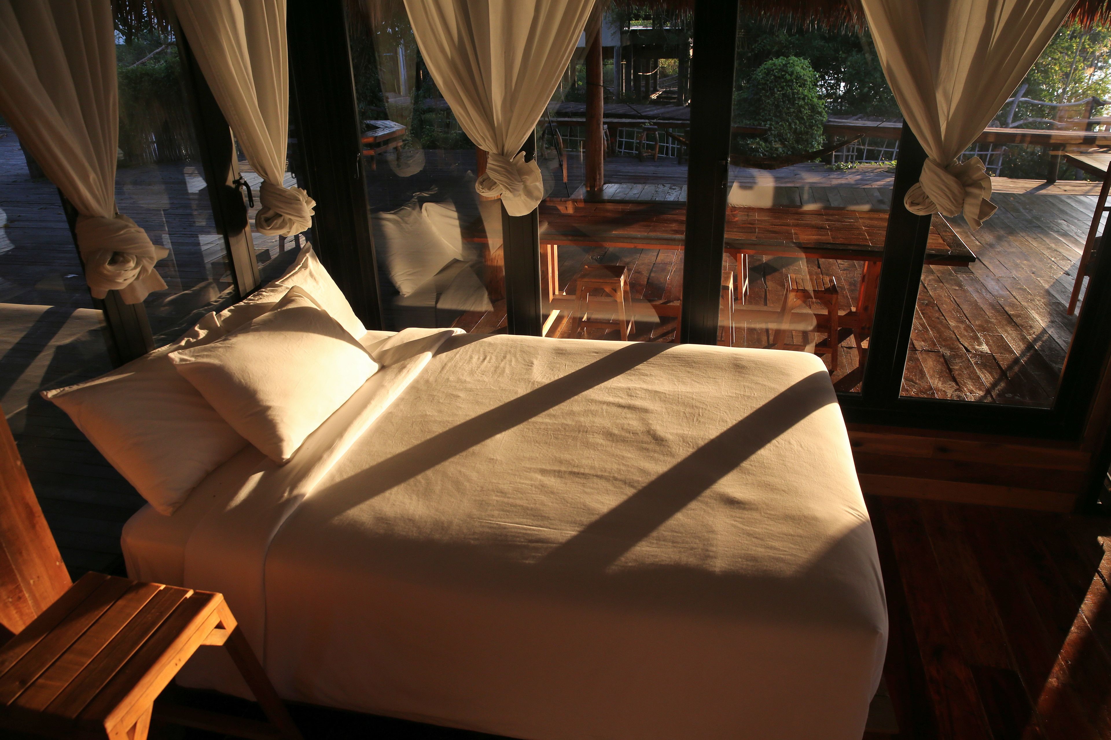 A bed with white linens in a room with large windows and wooden flooring.