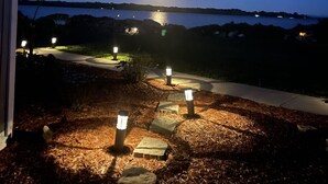 Une vue nocturne d’un espace paysager avec un sentier menant à un plan d’eau, illuminé par des éclairages au sol.