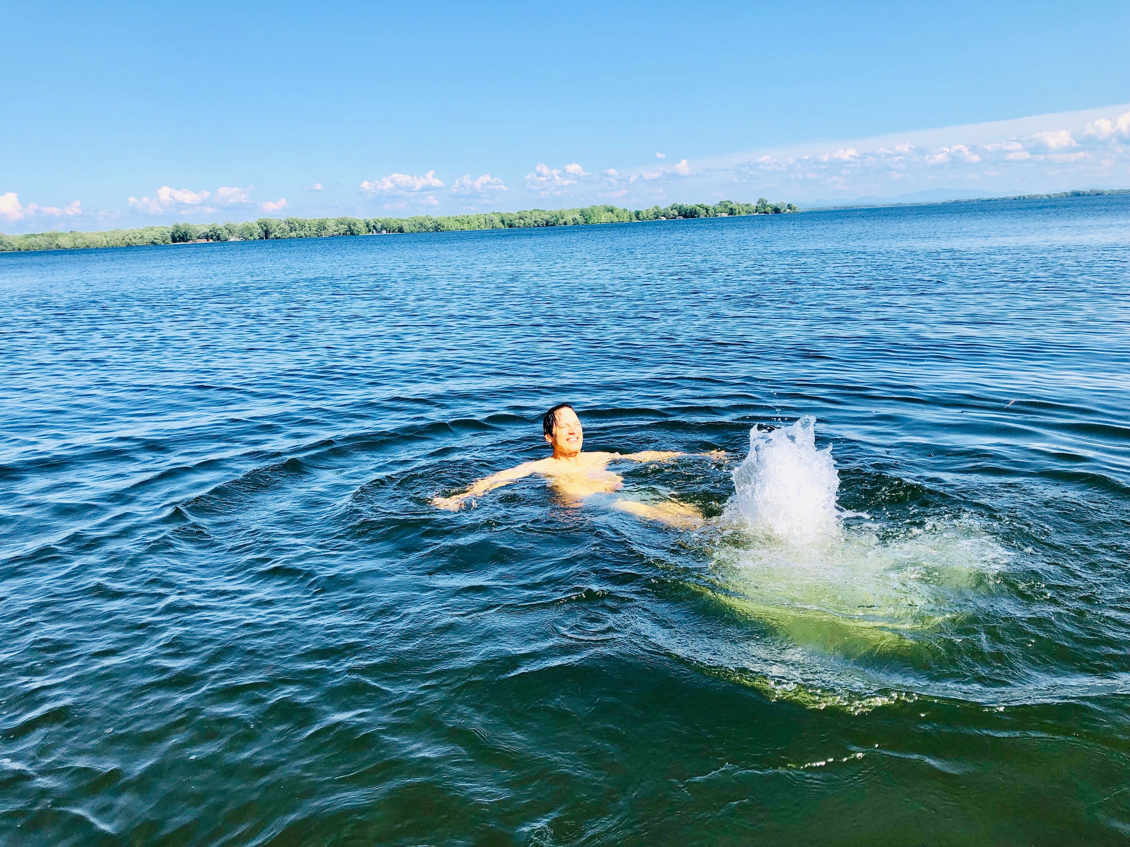 Une personne nage dans une étendue d’eau importante, avec une côte et des arbres visibles au loin.