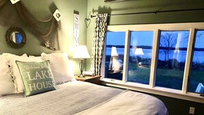 Une chambre à coucher avec un lit, une table de chevet, une lampe, une fenêtre donnant sur l’extérieur et un coussin orné de la mention « Bienvenue dans notre maison au bord du lac ».