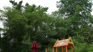 A playground with a slide and climbing structures, surrounded by a fence. There are chairs and a table with umbrellas in the foreground. - Bosco Homestay (Kota Kinabalu)
