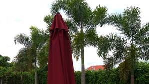 Una zona patio con due poltrone di legno e un ombrello rosso, circondata da palme e un pavimento piastrellato.