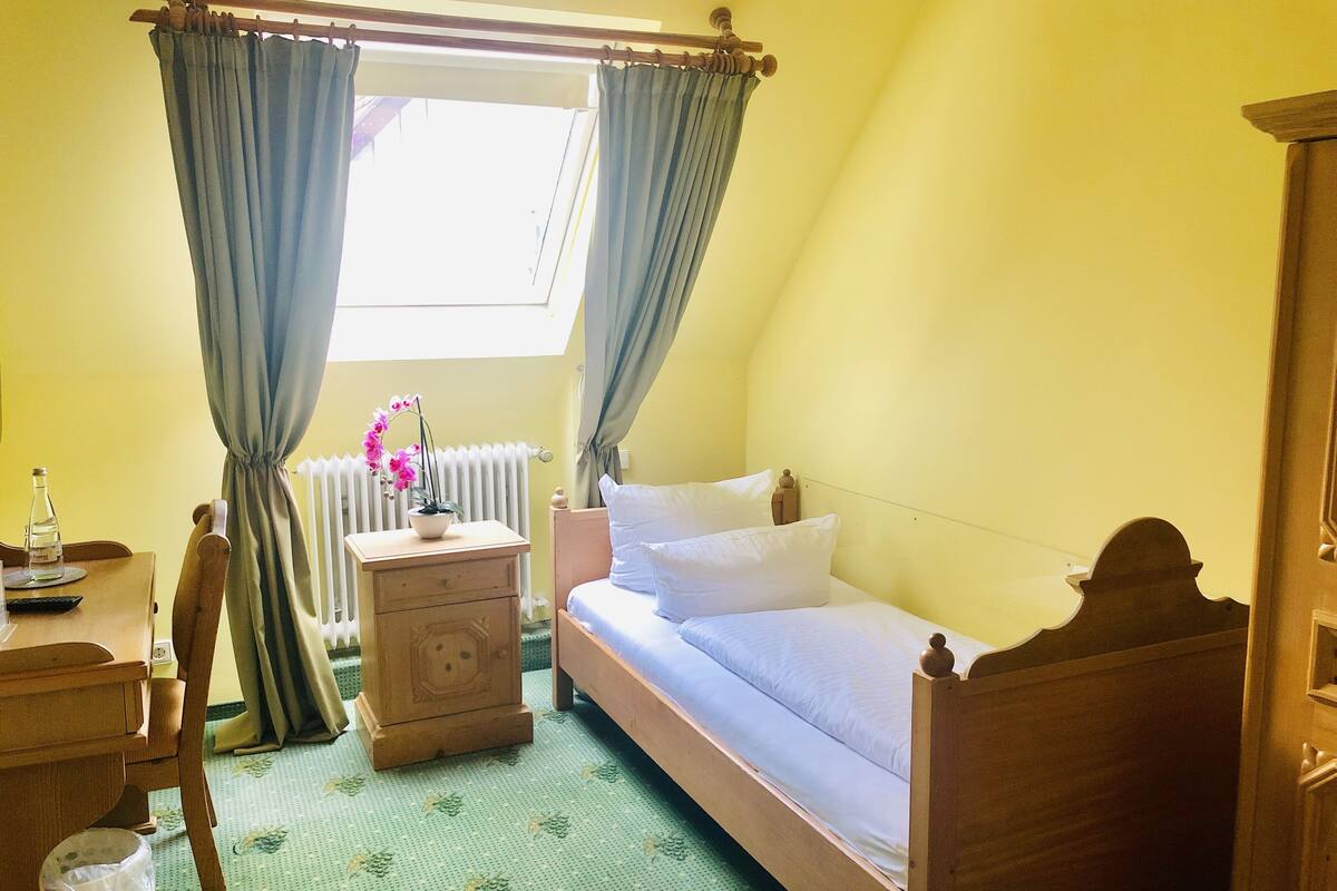 A bedroom with a wooden bed, a bedside table, a chair, a radiator, and a window with curtains.
