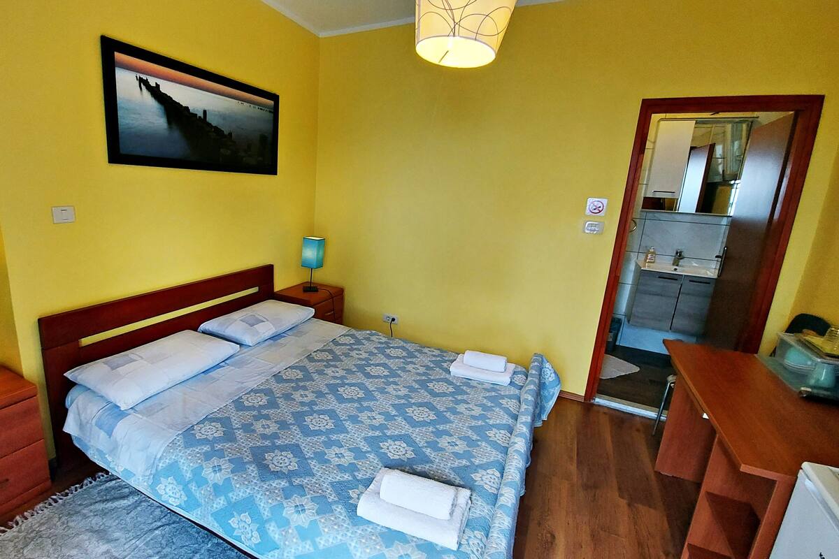 Ein Schlafzimmer mit Bett, Nachttischen, Schreibtisch, Stuhl, Lampe und Spiegel. Durch eine offene Tür ist ein Badezimmer zu sehen.