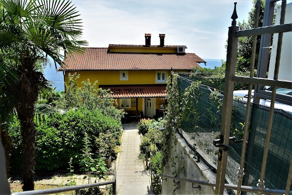 A yellow building with a red-tiled roof, a paved pathway leading to the entrance, and a view of the sea.