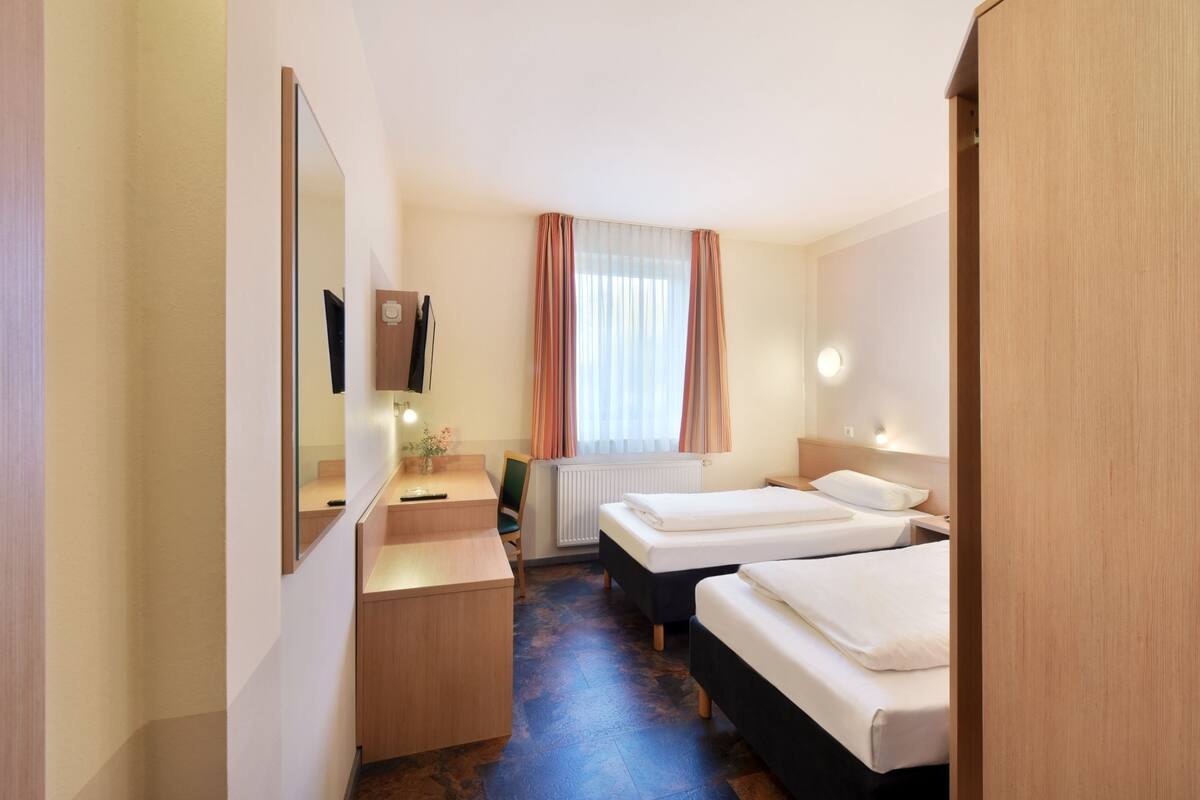 A hotel room with two beds, a desk, a chair, a mirror, a window with curtains, and a radiator.