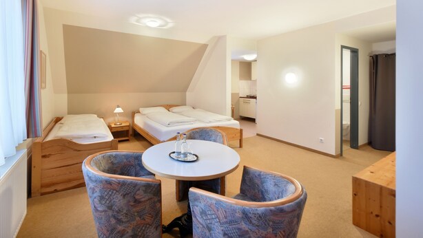 Habitación de hotel con dos camas individuales, una mesa redonda con sillas, un escritorio pequeño y una kitchenette al fondo.