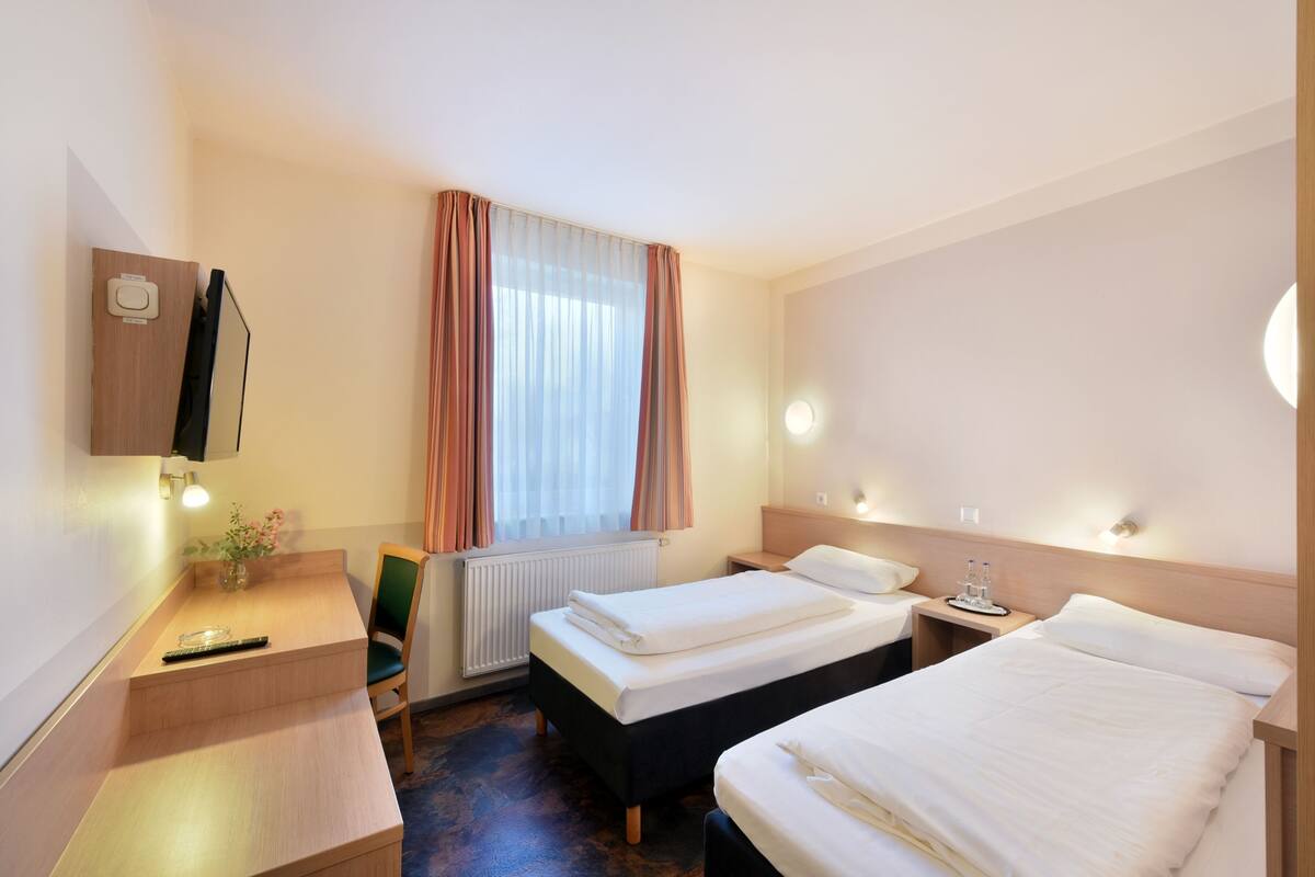 A hotel room with two beds, a desk with a chair, a small table with a vase, a radiator, and a window with curtains.