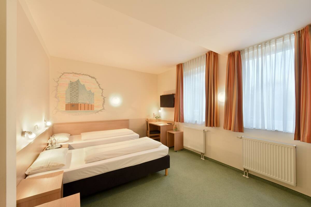 A hotel room with two beds, a desk, a chair, a radiator, and a window with curtains.