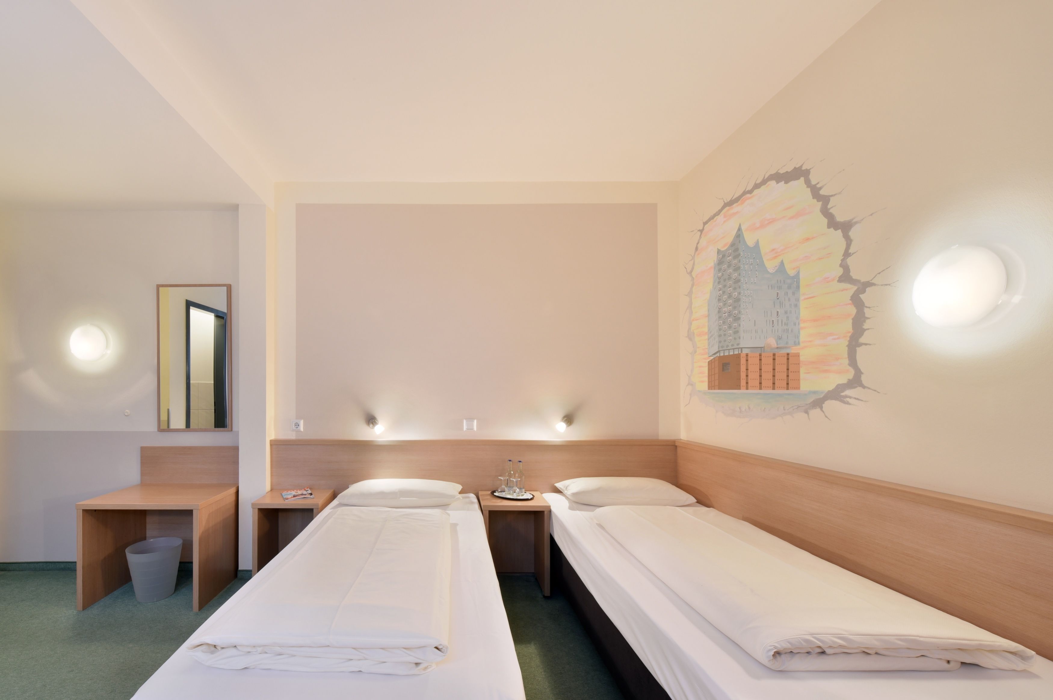 Habitación de hotel con dos camas individuales, una mesita con un cesto de basura y una pared decorada con un mural.