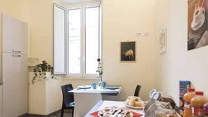 Un petit coin repas avec une table, des chaises et une fenêtre. On peut voir de la nourriture sur la table, une plante et une affiche accrochée au mur.