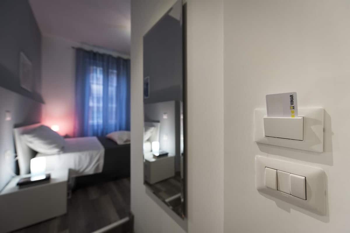 A modern hotel room with a bed, bedside table, and a wall-mounted control panel.