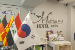 Zona de recepción de un hotel, con banderas, un cartel que dice "Maestro Hotel" y folletos informativos.