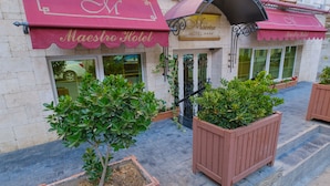 A hotel with a red awning and a sign that reads "Maestro Hotel." There are potted plants and a paved walkway in front. - Maestro Hotel (Baku)