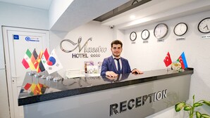 A hotel reception desk with a "Reception" sign, flags, clocks showing different time zones, and a potted plant. - Maestro Hotel (Baku)