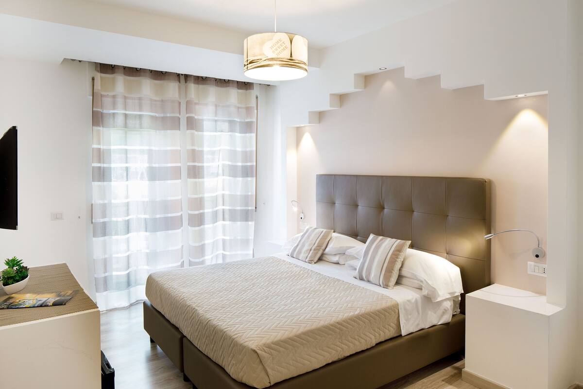 Ein modernes Schlafzimmer mit einem großen Bett, einem Nachttisch, einer Lampe und einem Fenster mit transparenten Vorhängen.