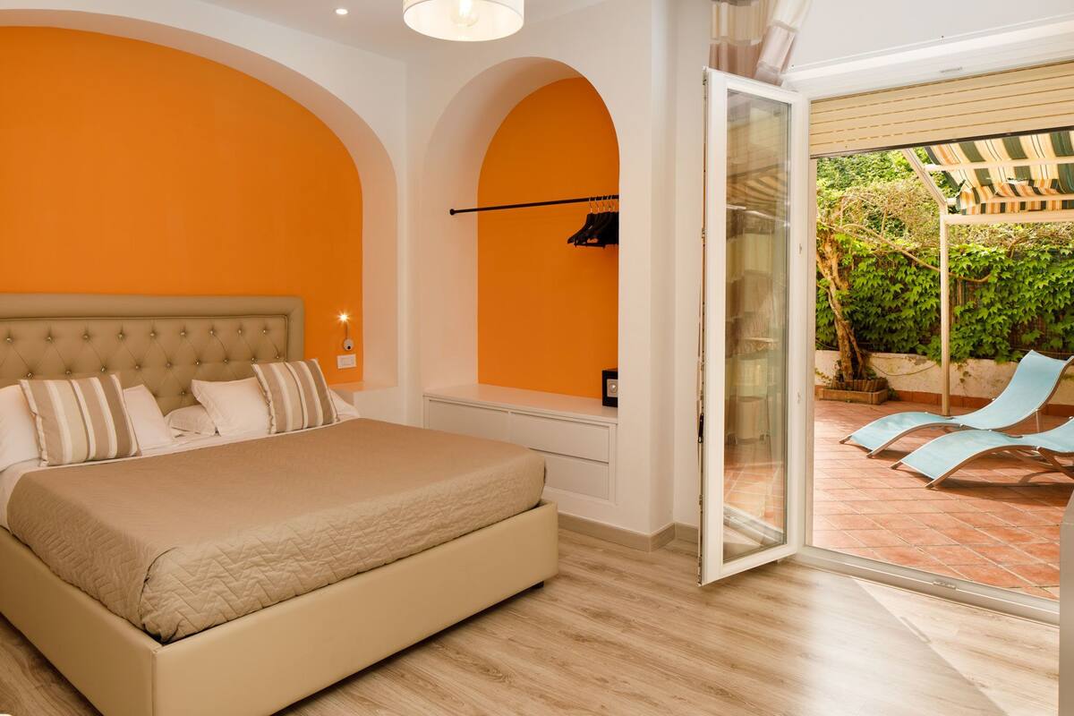 Ein Schlafzimmer mit Bett, Nachttisch und Blick auf einen Außenbereich mit Pergola und Liegestühlen.