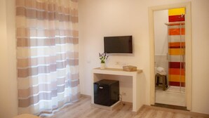 Une chambre moderne dotée d’un téléviseur à écran plat fixé au mur, d’une petite table en bois ornée d’un vase de fleurs, d’un meuble de rangement noir et compact, et d’un espace douche avec un rideau rayé aux couleurs vives.