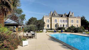 Un edificio grande y elegante con piscina, rodeado de exuberante vegetación y mobiliario de exterior.