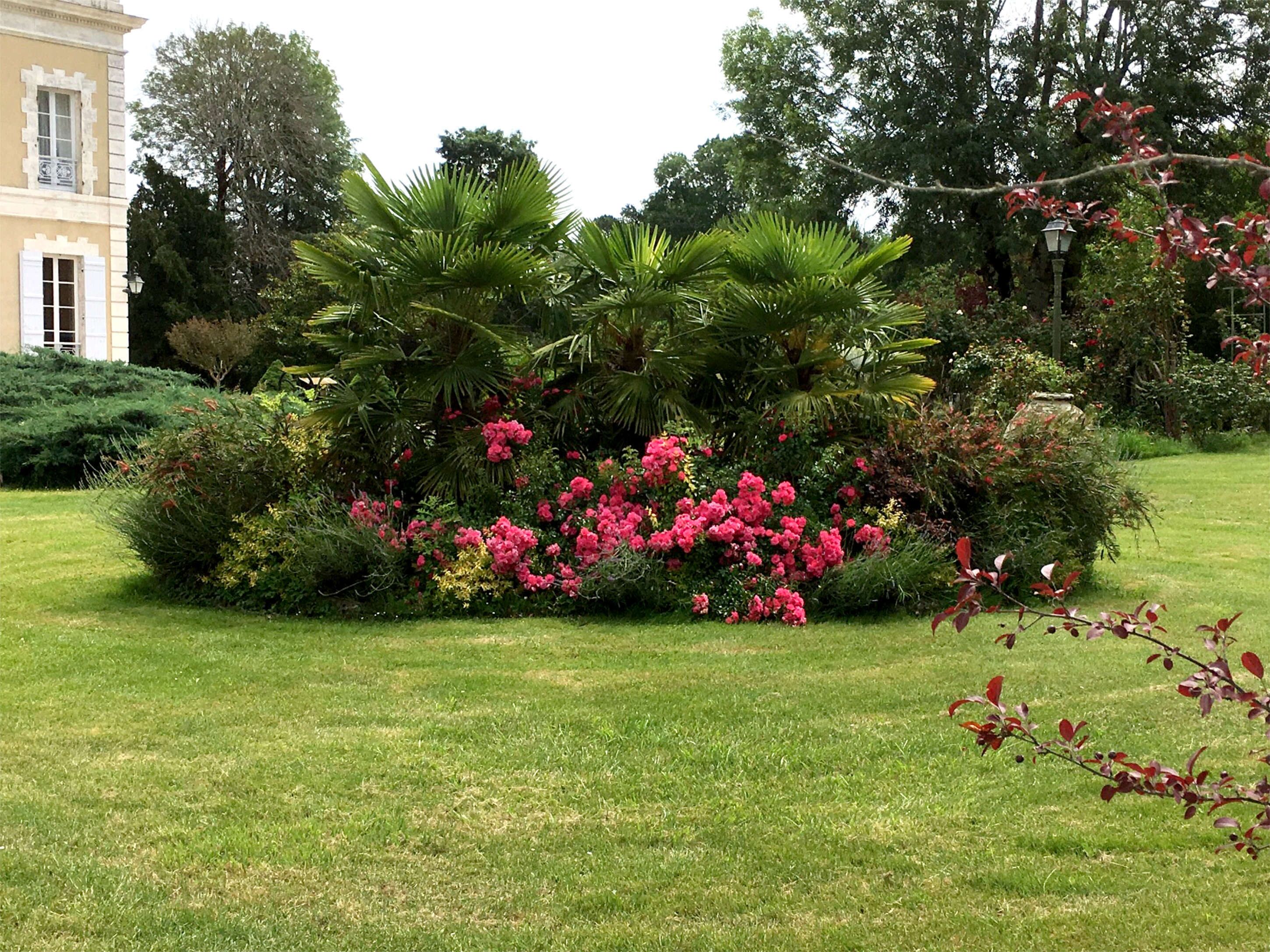 Un jardín bien cuidado con diversas plantas y flores, un césped y un edificio con arquitectura clásica al fondo.