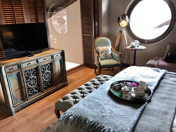 A cozy room with a television on a wooden cabinet, a round window, a chair with a cushion, and a tray with cups and a teapot on a bed. - Regnum Golf Country 3 Bedroom 2 (Bodrum)