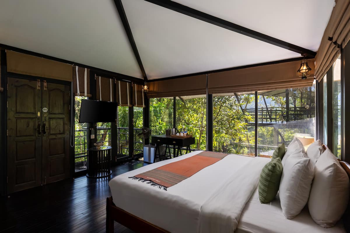 A bedroom with a large bed, a desk, and a chair, with a view of trees through large windows.
