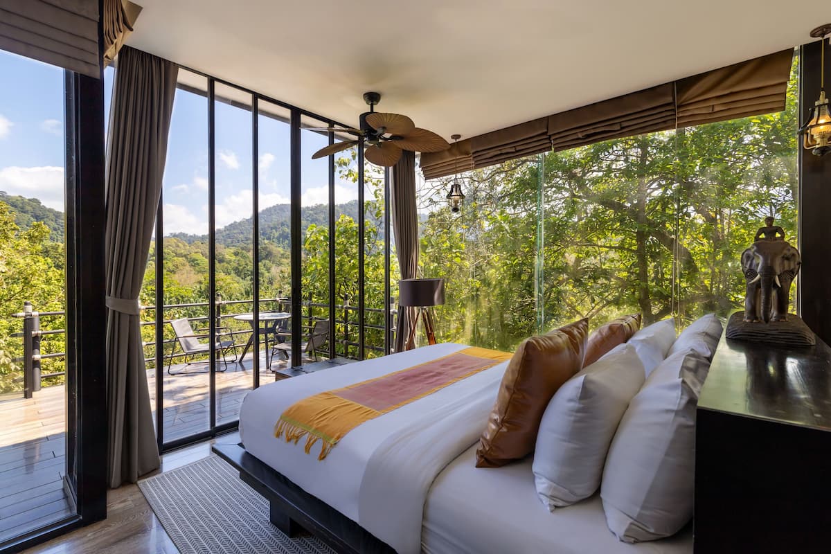A bedroom with a large bed, a ceiling fan, and a view of a lush forest through large windows.