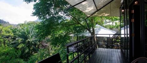 A wooden deck with a canopy, overlooking a lush green landscape with trees and a white tent in the background.