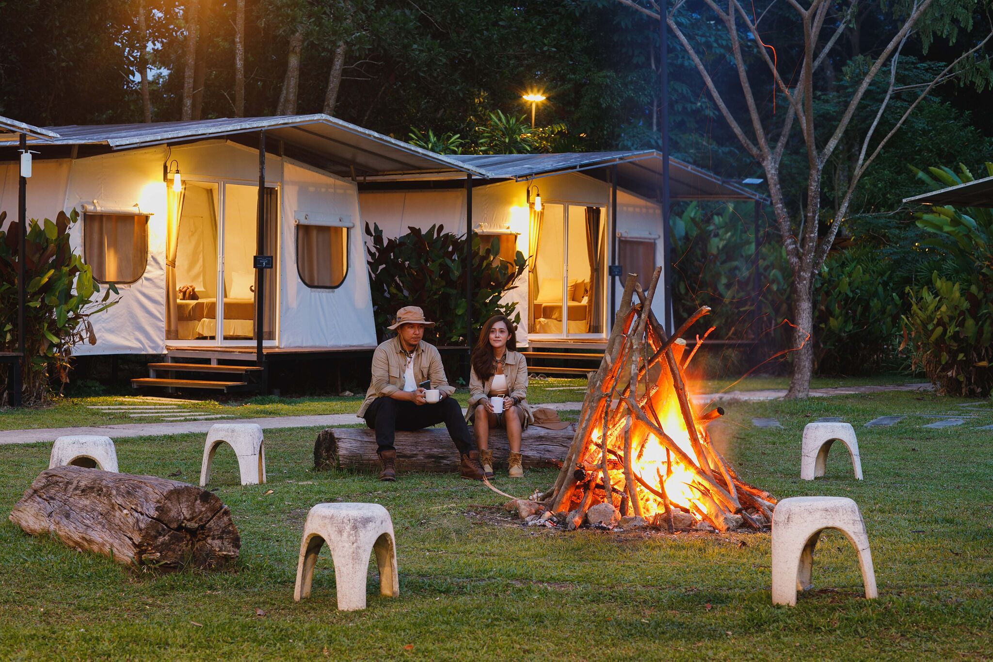 Ein Campingplatz mit Feuerstelle, zwei Personen sitzen auf Bänken, und ein Wohnwagen mit Veranda.