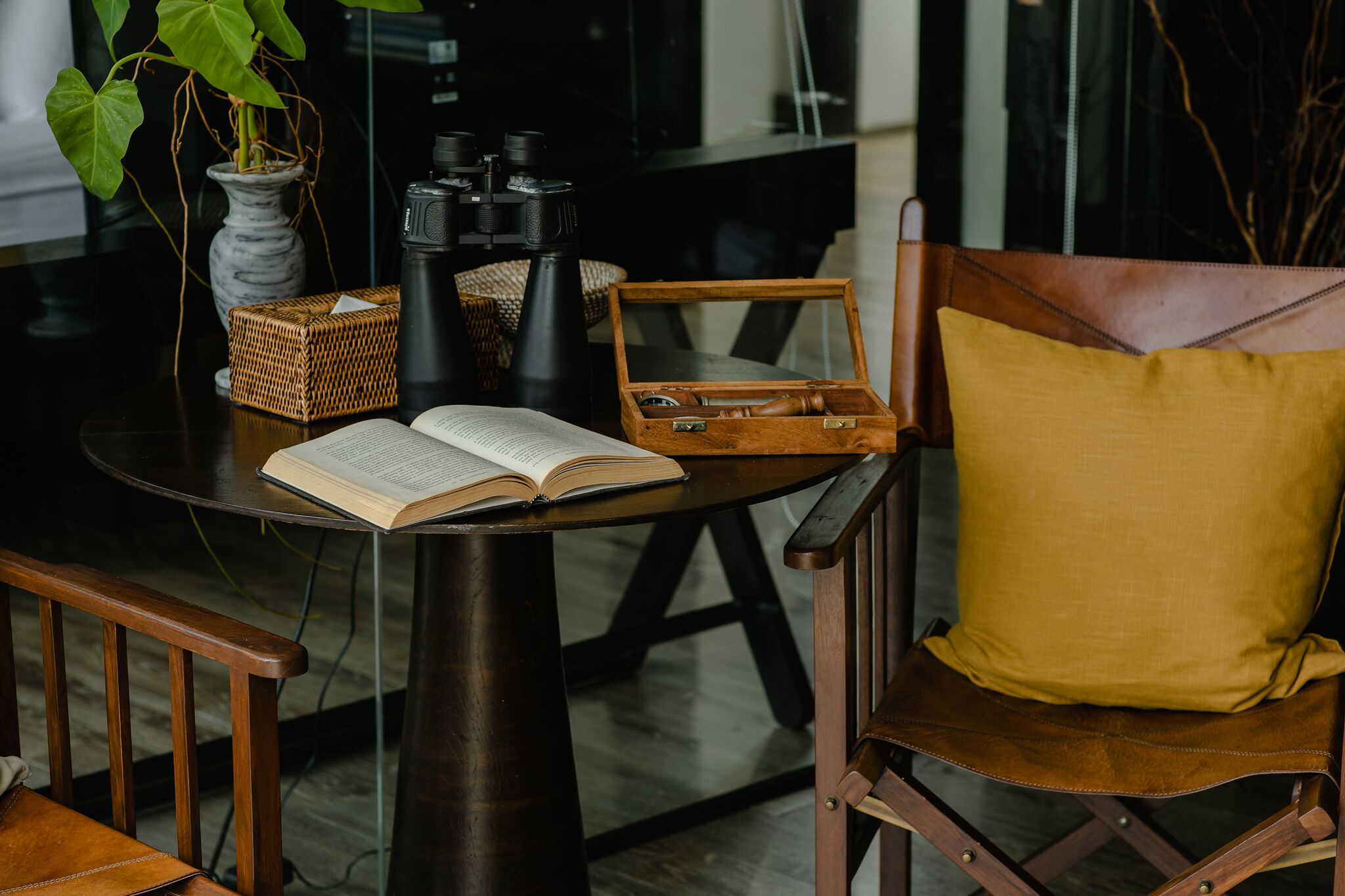 A table with an open book, a camera, binoculars, a wicker box, and a wooden box with a cushion.